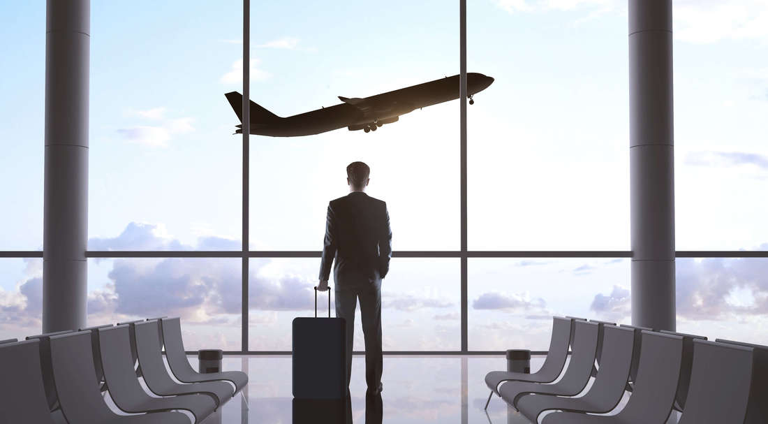 A man on a suit holding its suit case, standing in an airport watching an airplane flying.